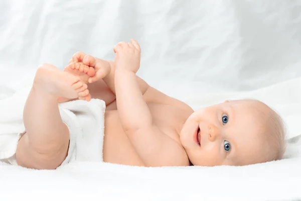 Roztomilé Šťastné Dítě Modrýma Očima Ležící Bílé Posteli Držící Nohy — Stock fotografie