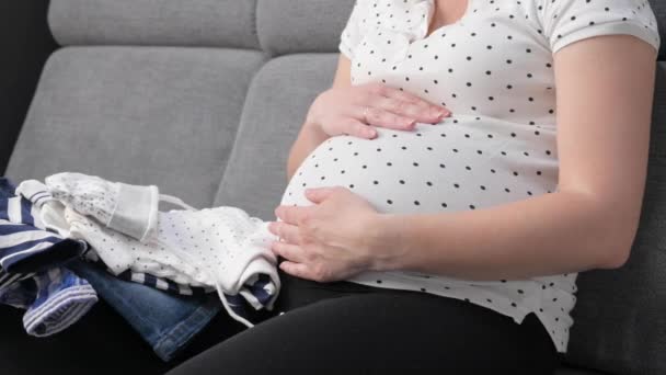 Happy Pregnant Belly Woman Prepares Baby Clothes While Sitting Sofa — Vídeos de Stock