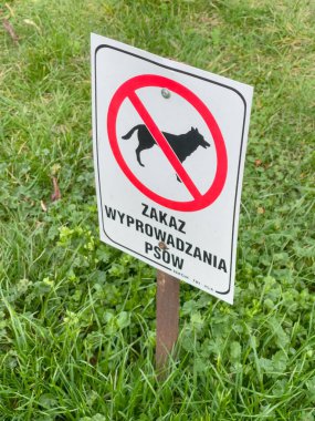 White metal Sign with inscription prohibiting dog walking and ban on defecation in park with green grass. No dogs sign. Inscription in polish.