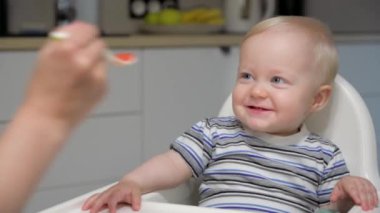 Anne oğlunu kaşıkla bebek mamasıyla besliyor. Çocuk yüksek sandalyede oturarak yulaf lapası yiyor. Annem mutfakta meyveli püreyi besler..