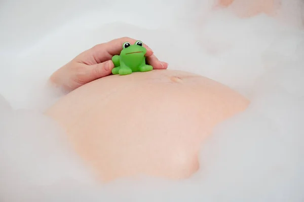 緑のカエルのおもちゃで妊娠中の腹 クローズアップ バブルバスを持っている妊婦 — ストック写真