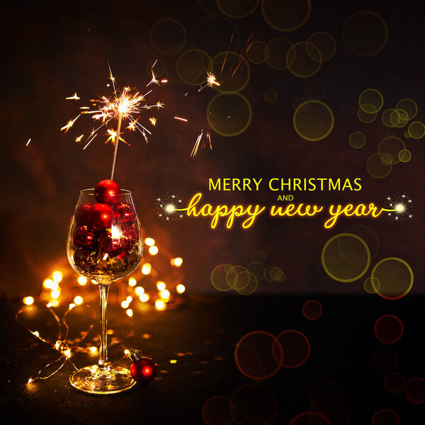 Merry Christmas and Happy New Year 2023. Wine glass with red christmas balls and burning sparkler, festive night, dark background, bokeh, selective focus. greeting card