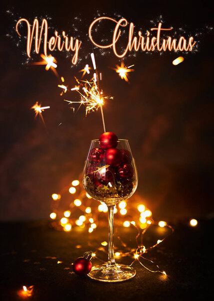 Merry Christmas 2023. Wine glass with red christmas balls and burning sparkler, festive night, dark background, bokeh, selective focus. greeting card