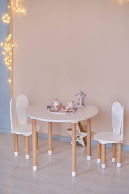 Small table with tea set and chairs with bunny ears in children's room interior, led lights. White and wooden colors. Scandi style