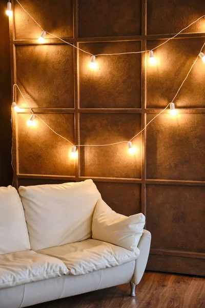 Empty comfy white couch, contemporary furniture inside of cozy living room interior with a garland of luminous lamps. Wooden wall, brown laminate. Real estate, loan or rent