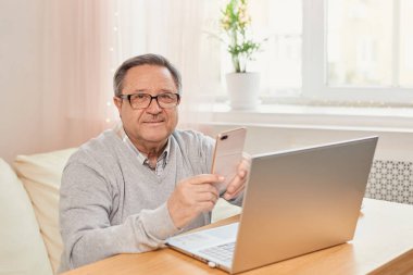 Cheerful senior man has phone conversation, chatting on the smartphone working with a laptop remotely from home.