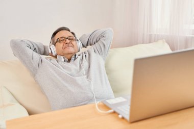 Rest And Relax Concept. Excited mature man sitting leaning back on couch listening to music, audio book or podcast, enjoying in headphones holding hands behind head.