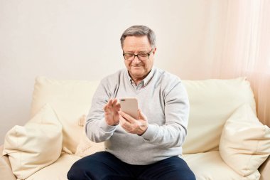 People And Technology. Portrait of smiling mature man using mobile phone, watching video or reading sms message, sitting on the couch in living room at home. Elderly man uses modern technology