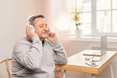 Senior man with closed eyes in living room wearing headphones listens favourite track, having nostalgic mood, enjoys songs of his youth, older generation using modern technology concept