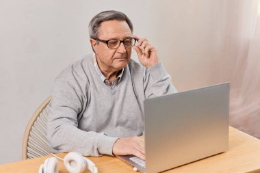 Senior man in casual clothing and eyeglasses using laptop and smiling while sitting at the desk at home