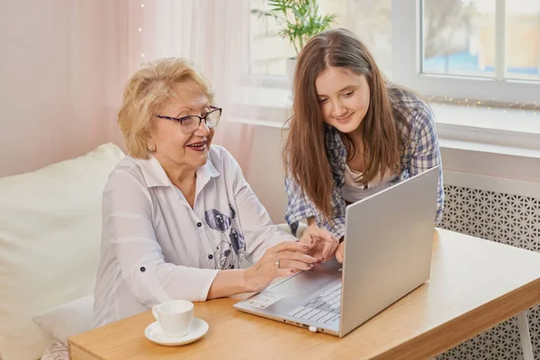 Happy grandchild and old grandmother sitting at home using laptop. Smiling cute girl and senior woman on laptop. Little granddaughter teach her beautiful granny how to use computer at home.