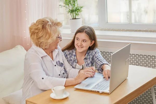 Happy grandchild and old grandmother sitting at home using laptop. Smiling cute girl and senior woman on laptop. Little granddaughter teach her beautiful granny how to use computer at home.