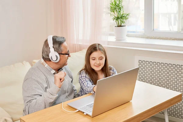 Granddaughter helping grandpa to make on line communication on laptop, listening to modern music, old man with his granddaughter using laptop making video call
