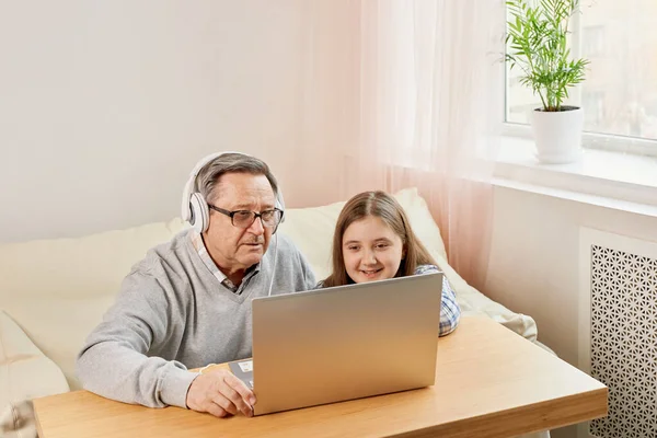 Granddaughter helping grandpa to make on line communication on laptop, listening to modern music, old man with his granddaughter using laptop making video call
