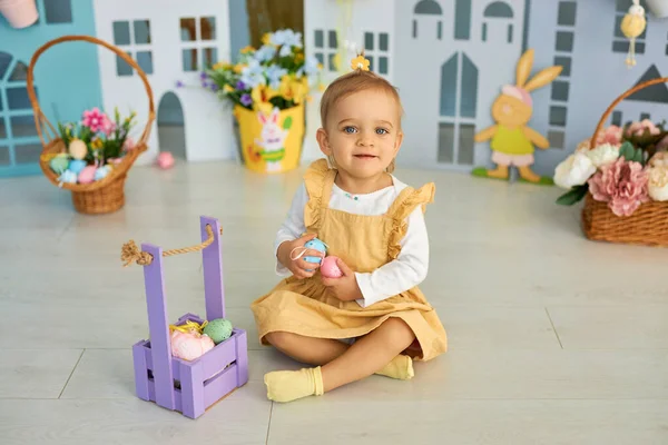 Baby girl celebrate Easter. Funny happy kid playing on Easter egg hunt. Family home decoration, colorful Easter eggs and flowers. Home decoration and flowers