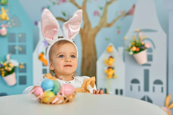 Baby girl celebrate Easter. Funny happy kid in bunny ears Sits by the table and playing with Easter eggs. Colorful Easter eggs and flowers. Home decoration and flowers