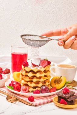 Ev yapımı geleneksel Belçika waffleları. Meyveli ve bir fincan kahveli. Seçici odaklı. Bir kadının eli gözlemelere şeker serpiyor.