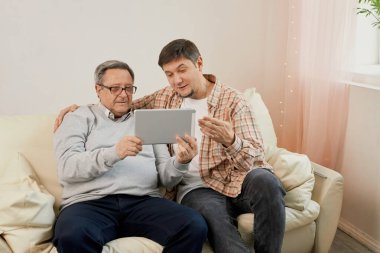 Öğretmen oğlu. Gülümseyen genç erkek sevgili olgun babaya bilgisayardaki uygulamayı açıklamaktan memnuniyet duyar. Yaşlı baba ve yetişkin çocuk, e-postaları kontrol eden web alışveriş siteleri için tablet kullanıyor