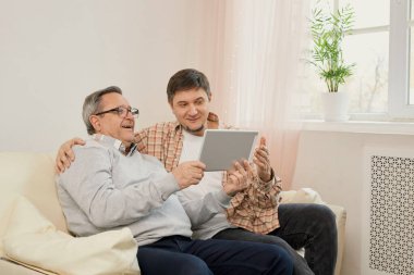 Öğretmen oğlu. Gülümseyen genç erkek sevgili olgun babaya bilgisayardaki uygulamayı açıklamaktan memnuniyet duyar. Yaşlı baba ve yetişkin çocuk, e-postaları kontrol eden web alışveriş siteleri için tablet kullanıyor
