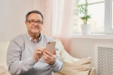 People And Technology. Portrait of smiling mature man using mobile phone, watching video or reading sms message, sitting on the couch in living room at home. Elderly man uses modern technology