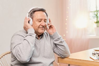 Senior man with closed eyes against a gray background wearing headphones listens music, having nostalgic mood, enjoys songs of his youth, older generation using modern technology concept, copy space