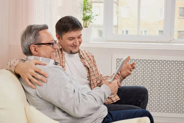 teacher son. Smiling young male glad to explain work online in app on laptop to beloved mature dad. Elderly father and adult kid use tablet for web shopping browsing sites checking email