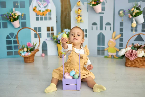 Baby girl celebrate Easter. Funny happy kid playing on Easter egg hunt. Family home decoration, colorful Easter eggs and flowers. Home decoration and flowers