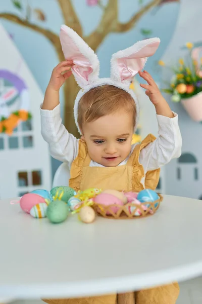 Baby girl celebrate Easter. Funny happy kid in bunny ears Sits by the table and playing with Easter eggs. Colorful Easter eggs and flowers. Home decoration and flowers