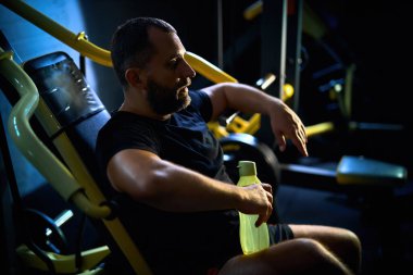 Antrenmanından memnun. Yakışıklı orta yaşlı bir adam elinde bir şişe suyla karanlık bir salonda oturuyor, egzersizden sonra dinleniyor.