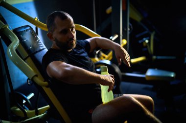 Antrenmanından memnun. Yakışıklı orta yaşlı bir adam elinde bir şişe suyla karanlık bir salonda oturuyor, egzersizden sonra dinleniyor.