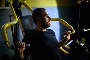 Orta yaşlı bir adam spor salonunda göğüs presi makinesinde çalışıyor, ağırlık kaldırıyor, kollarını ve gövde kaslarını geliştiriyor.