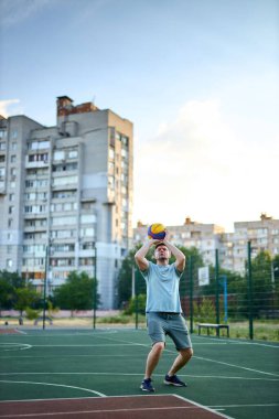 Güneşli bir günde orta yaşlı bir adam şehir kortunda basketbol oynuyor. Dışarıda spor yapan güçlü bir adam. Arka planda konut binaları ve ağaçlar var. Spor, aktif yaşam tarzı konsepti