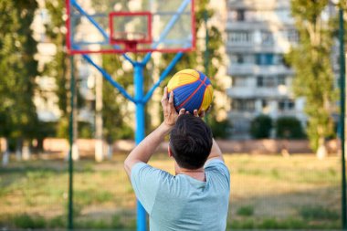 Güneşli bir günde orta yaşlı bir adam şehir kortunda basketbol oynuyor. Dışarıda spor yapan güçlü bir adam. Arka planda konut binaları ve ağaçlar var. Spor, aktif yaşam tarzı konsepti