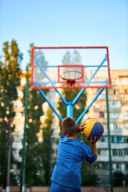Oyun sahasında basketbol oynayan neşeli, genç bir çocuk. Çocuk şehir binalarının arka planına karşı Red Court maçında atış yapıyor. Aktif yaşam tarzı, eğitim, profesyonel spor