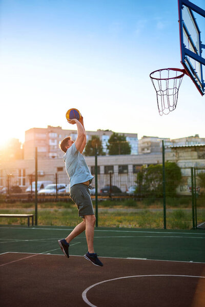 Middle aged man playing basketball on city court on sunny day. Strong man doing sports outside. Residential buildings and trees in the background. Sport, active lifestyle concept