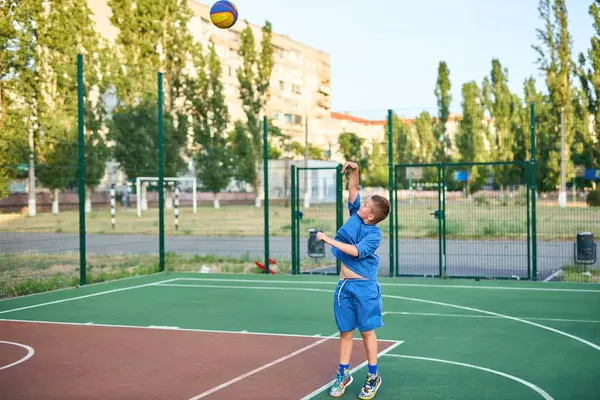 Stock image Joyful young sport boy kid playing basketball on playground. Boy performs shot at red court against the backdrop of urban residential buildings. Active lifestyle, training, professional sports