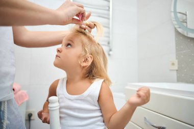 Mutlu bir aile. Anne küçük kızının saçını tarıyor ve banyoda saçını yapıyor. Neşeli ve duygusal bir çocuk ve mutlu bir anne.