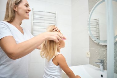 Mutlu bir aile. Anne küçük kızının saçını tarıyor ve banyoda saçını yapıyor. Neşeli ve duygusal bir çocuk ve mutlu bir anne.