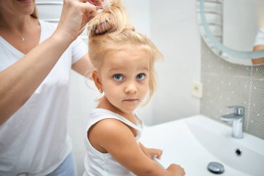 Mutlu bir aile. Anne küçük kızının saçını tarıyor ve banyoda saçını yapıyor. Neşeli ve duygusal bir çocuk ve mutlu bir anne.