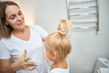 Mutlu bir aile. Anne küçük kızının saçını tarıyor ve banyoda saçını yapıyor. Neşeli ve duygusal bir çocuk ve mutlu bir anne.