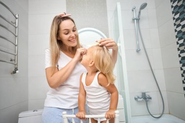 Genç anne ve kızı banyoda bigudi yapıyor, mutlu bir şekilde gülümsüyorlar. Banyo odasında bakım konsepti