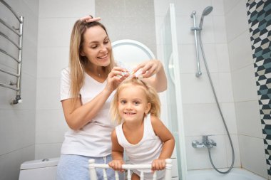 Genç anne ve kızı banyoda bigudi yapıyor, mutlu bir şekilde gülümsüyorlar. Banyo odasında bakım konsepti