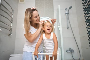 Genç anne ve kızı banyoda bigudi yapıyor, mutlu bir şekilde gülümsüyorlar. Banyo odasında bakım konsepti