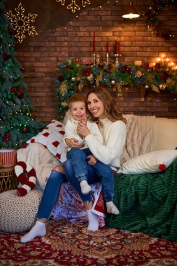 Mutlu anne ve küçük çocuk Noel tatili boyunca evde rahat vakit geçirirken Noel ağacının yanındaki kanepede birlikte oturuyorlar. Kapalı etkinlikler.