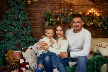 Mutlu aile Noel ağacının yanında sımsıkı sarılıyor ve güzel bir evde Noel arifesinde neşeyle gülümsüyor.