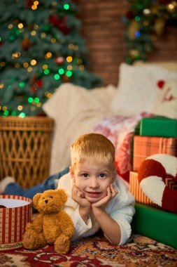 Üç yaşındaki mutlu çocuk evde çelenk ve hediye kutularıyla Noel ağacının yanında yerde yatıyor. Noel ve Yeni Yıl gelenekleri.