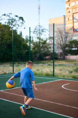Spor ve basketbol. Mavi eşofmanlı çocuk basketbol oynuyor, topa önderlik ediyor. Arka planda yerleşim alanı var. Boşluğu kopyala