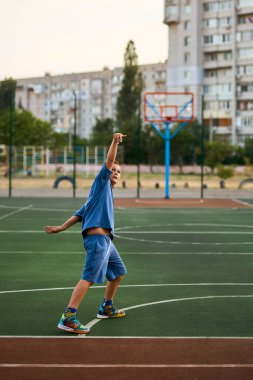 Spor ve basketbol. Mavi eşofmanlı bir çocuk dışarıda oynarken basketbol topunu potaya atıyor. Arka planda yerleşim alanı var. Spor konsepti