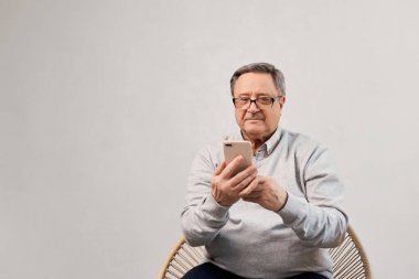 Üst düzey bir adam sandalyede otururken akıllı telefonunu dikkatle kullanıyor. Teknoloji, iletişim ve kıdemli yaşam tarzı konsepti