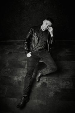 Deri ceketli, siyah gömlekli, kot pantolonlu, düşünen ya da dinleyen şık bir adamın tek renkli tüm vücut pozu. Dekor, dokulu duvar ve zemin ile kapalı gibi görünüyor.